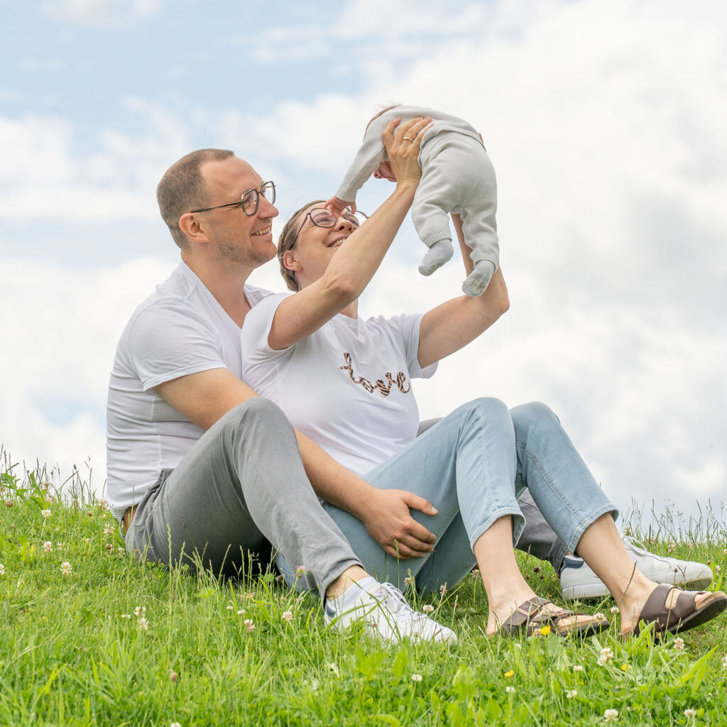 Junge Eltern verbringen eine schöne Zeit mit ihrem Baby. Der Vater umarmt tdie Mutter von hinten. Beide schauen dem Baby ins Gesicht.